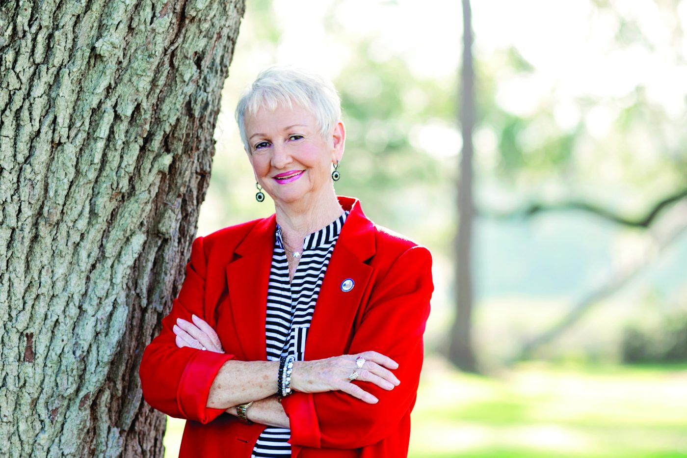 Judy Wise smiling with her arms crossed, wearing a red blazer and leaning against an oak tree